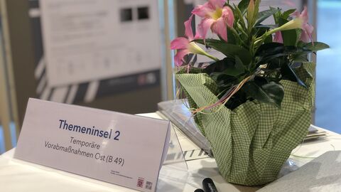 Stehtisch bei der Infomesse: Darauf befindet sich ein Schild mit der Aufschrift "Themeninsel 1 - temporäre Vorabmaßnahmen Ost", daneben eine kleine Topfflanze mit rosa Blüten und Informationsmaterial