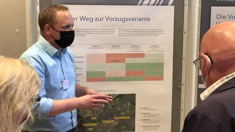Besucher bei der Infomesse Personen erläutern Besuchern den Weg zur Vorzugsvariante anhand eines Infoplakates welches an einer Pinnwand aufgehängt ist